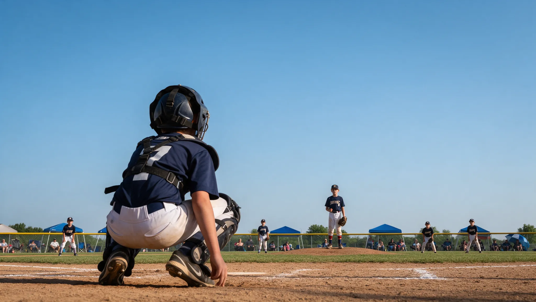 A youth catcher crouched behind home plate at a weekend tournament, the pitcher and parents' tents beyond
